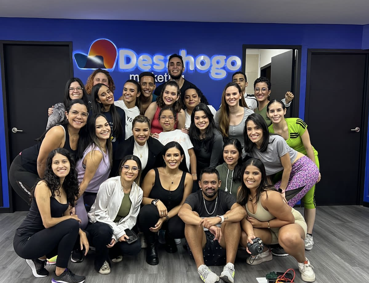 Gran grupo de mujeres y hombres diversos con ropa deportiva, posando en un estudio de baile con el logotipo de Deshogo.